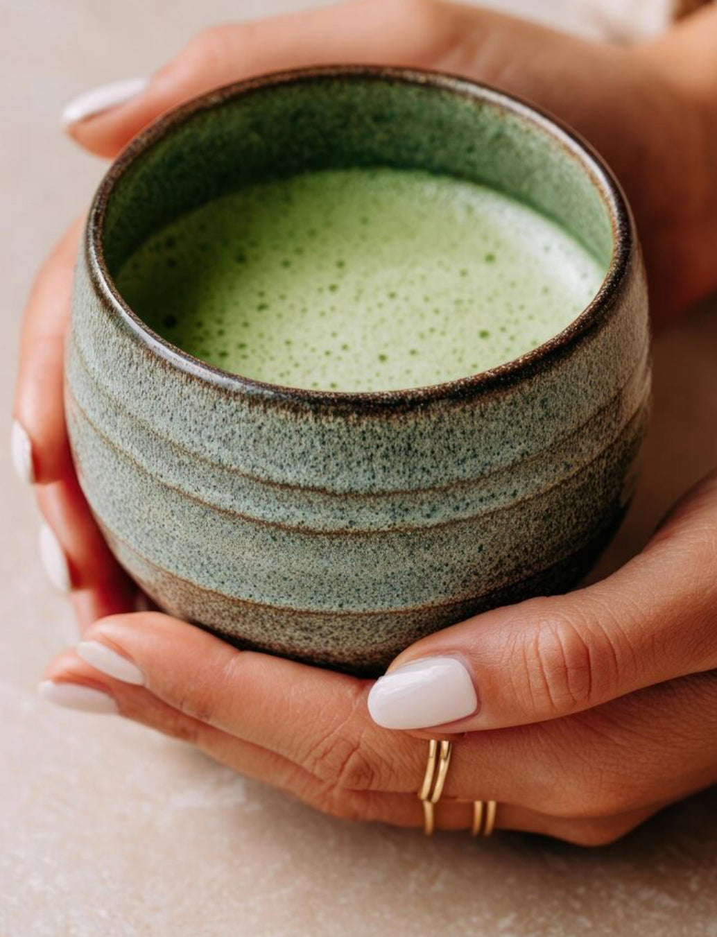 Mise en scène d'un bol de matcha prêt à déguster et mousseux. Gros plan sur des mains féminine tenant le bol, avec des ongles manucuré en blanc.