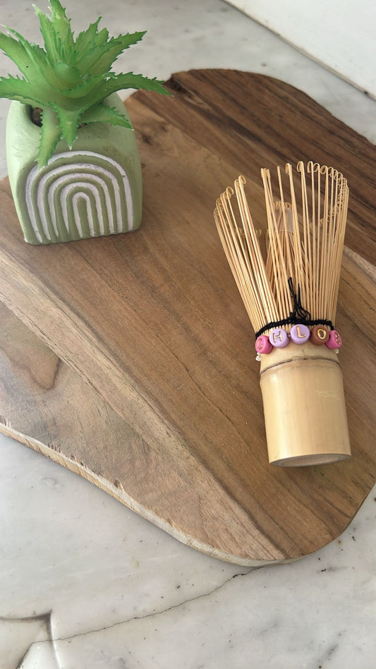Présentation d'un fouet chasen personnalisé sur une planche en bois avec une petite plante à gauche.
Le fouet est personnalisé avec le prénom Chloé grâce à des petites perles colorées avec des lettres gravés.