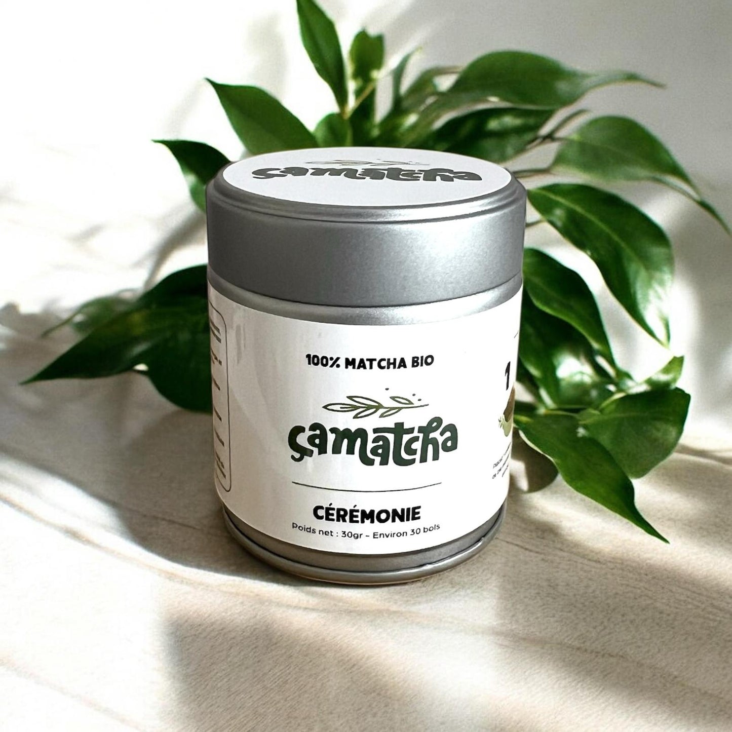 Présentation d'un pot de poudre matcha cérémonie ça matcha. Les feuilles de la plante derrière font ressortir les couleurs du logo présent sur l'étiquette.