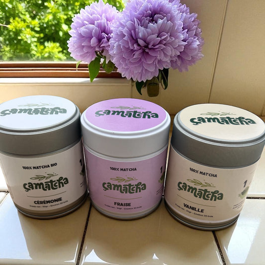 Présentation des 3 pots de poudre de matcha disponible dans la boutique: cérémonie, arôme fraise et arôme vanille. Ils sont posés sur le carrelage d'une cuisine avec une fleur violette au dessus, proche d'un fenêtre.