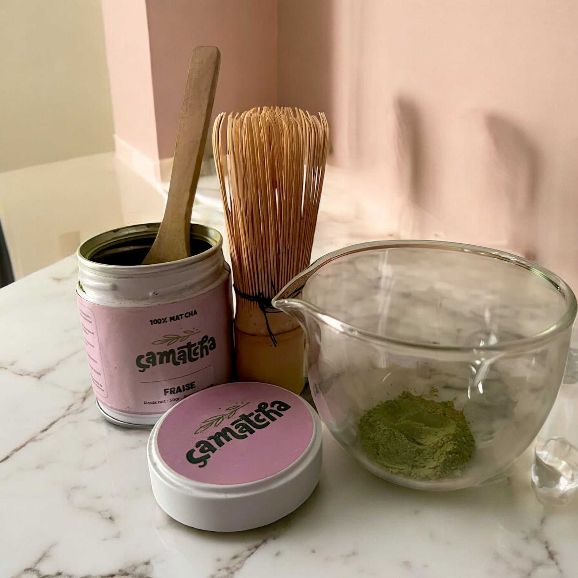 Set global de préparation avec un pot de poudre de matcha arôme fraise. On retrouve un bol avec un peu de poudre, un fouet chasen t une cuillère en bois posé dans un pot de poudre de matcha fraise ouvert.