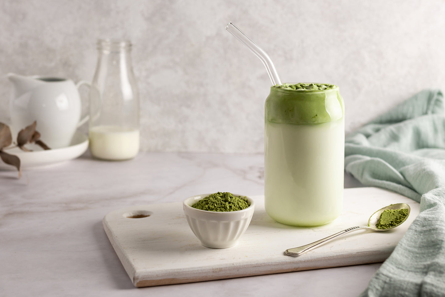 Présentation d'un grand verre de matcha version latte. On voit le verre avec une paille également en verre. A son côté, une bouteille de lait en verre, un bol remplis de poudre matcha ainsi qu'une cuillère en acier.