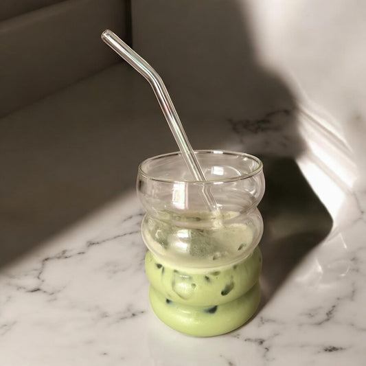 Présentation d'un verre à matcha design en verre, effet ondulé. Avec une paille en verre. A l'intérieur du verre se trouve un matcha latte cérémonie